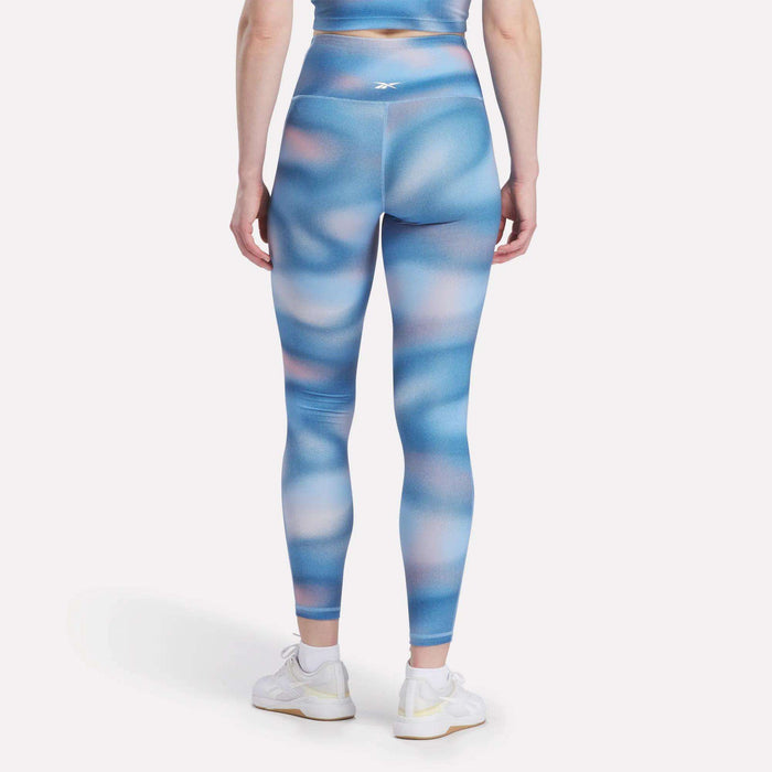 A woman faces away, wearing Reebok Women's Fluid Print High-Rise Tights with a matching top and white sneakers, all set against a plain white background.