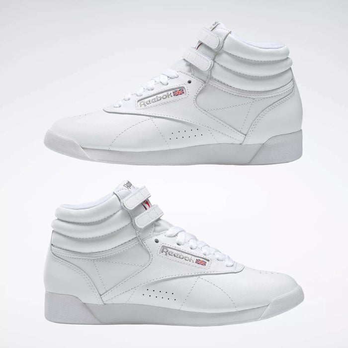 Two Reebok Women's Freestyle Hi Shoes in white are shown side by side on a light background, highlighting both outer and inner sides. Features include visible laces, velcro straps, and classic Reebok logos—ideal for women's fitness.