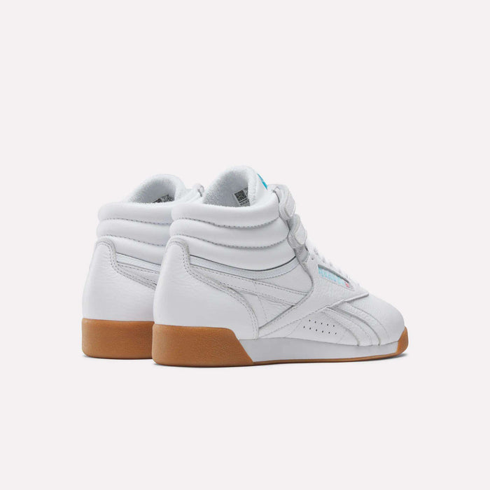 A pair of Reebok Women's Freestyle Hi Shoes in white with light brown rubber soles and padded ankles, inspired by the 80s fitness classic, is shown against a plain white background facing away from the viewer.