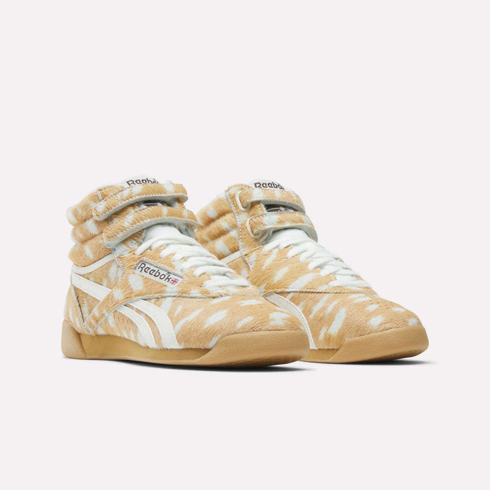Two Reebok Women's Freestyle Hi Shoes with tan and white faux fur, white laces, and gum soles are displayed on a white background. The Reebok logo appears on the side and tongue, evoking an 80s fitness classic style.