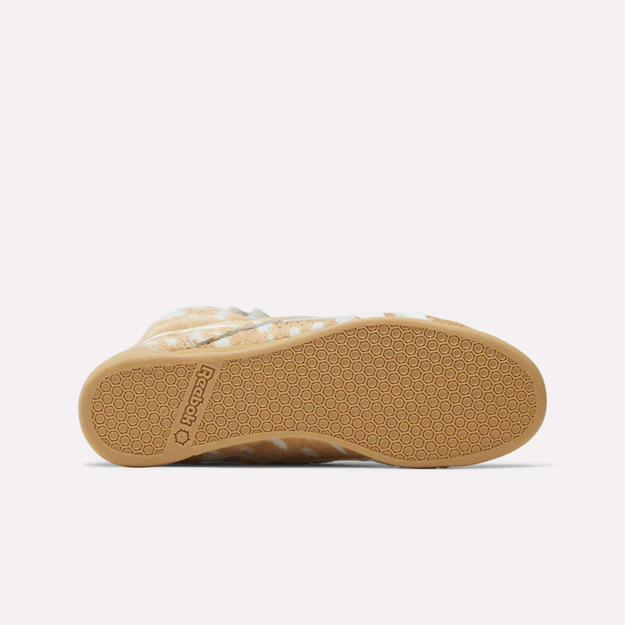 A bottom view of the Reebok Women's Freestyle Hi Shoes highlights a light brown, textured rubber sole with a honeycomb pattern and logo at the heel. The tan and white upper nods to classic 80s Reebok fitness high tops.