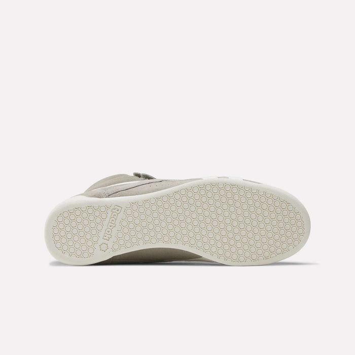 The image shows the bottom view of a beige Women's F/S Hi Shoe by Reebok, with a white sole featuring a hexagonal pattern and the Reebok name embossed near the heel, reflecting popular high top sneaker design trends.