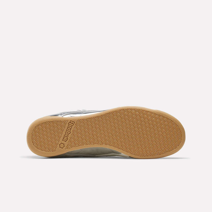 The image displays the bottom view of the Reebok Women’s Freestyle Hi Shoes, showcasing a gum rubber sole with a distinctive honeycomb pattern and signature Reebok branding. The upper is crafted from premium light-colored leather, providing both style and comfort.