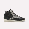 An Almostblack/Vintagechalk/Gold Women’s Freestyle Hi Zip Shoe by Reebok is displayed in side profile against a plain white background, featuring dual velcro straps, snakeskin-patterned accents, black laces, and an off-white sole. These retro-inspired sneakers blend iconic Reebok design with the comfort and durability of women’s leather footwear.