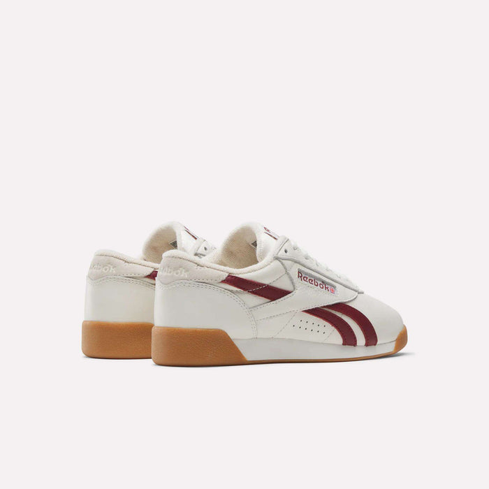 The Reebok Women's Freestyle Lo Shoes feature maroon stripes and logos, gum soles, beige accents, and white leather. They're displayed at an angle to show both the side and back views against a plain white background.