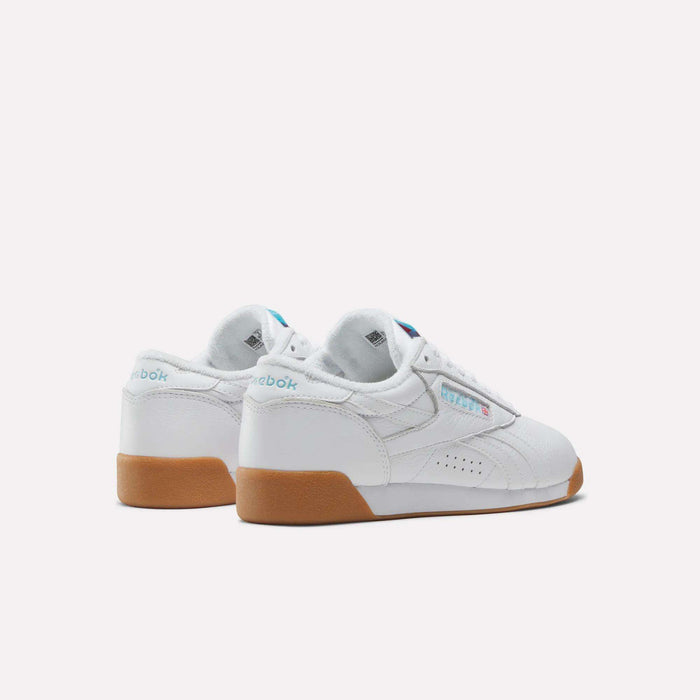 The Reebok Women's Freestyle Lo Shoes feature a white design with gum soles, light blue branding on the heel and side, and perforated details, shown against a plain white background.
