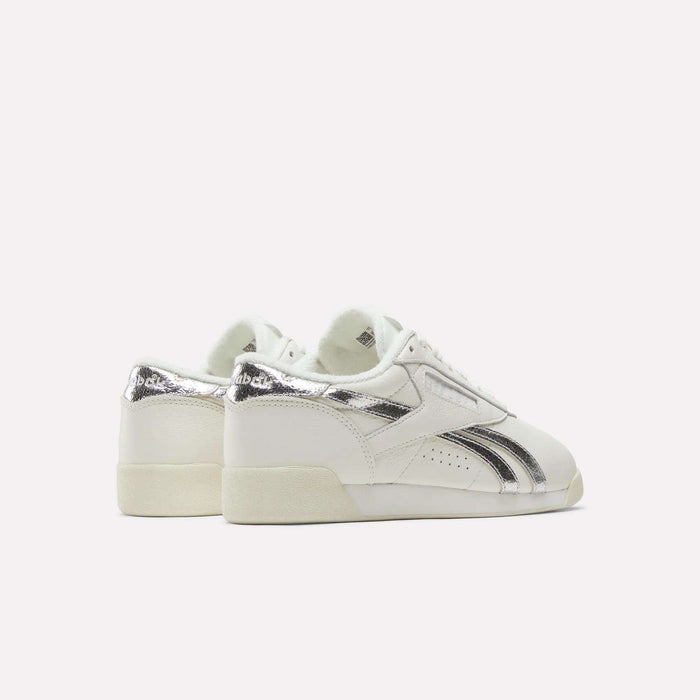 A pair of Reebok Women's Freestyle Lo Shoes featuring metallic accents on the heel and side stripes are displayed against a plain, light background with their heels angled slightly toward the viewer.