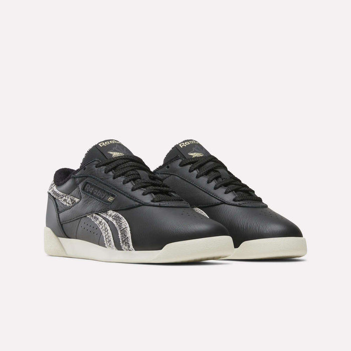 A pair of Reebok Women’s Freestyle Lo Shoes in black, showcasing off-white soles and gray snakeskin-patterned accents on the sides, are displayed against a plain white background.