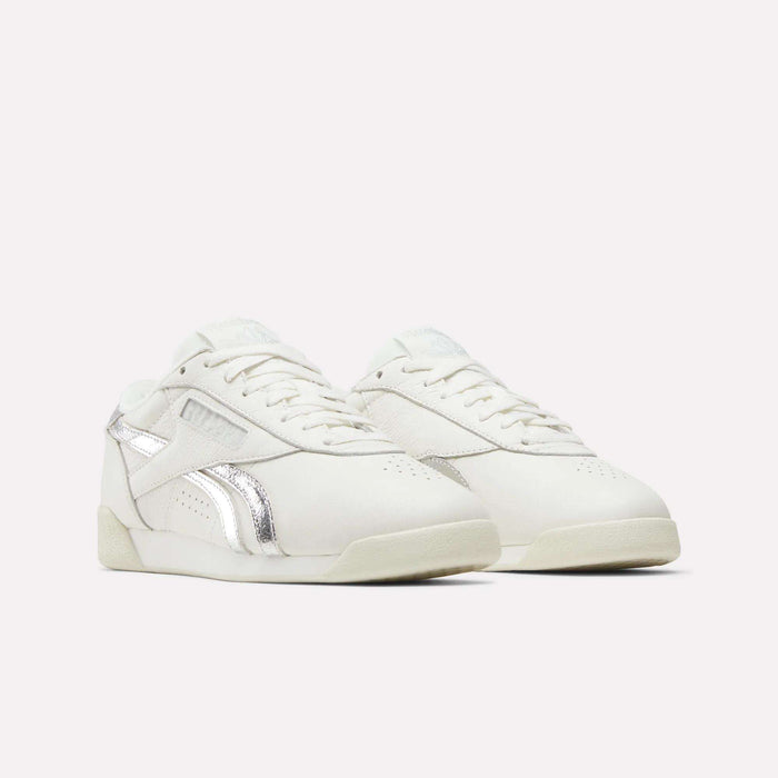 A pair of Reebok Women's Freestyle Lo Shoes with silver metallic accents is shown against a plain light background. These sneakers have a low-top design and sport a minimalistic style.