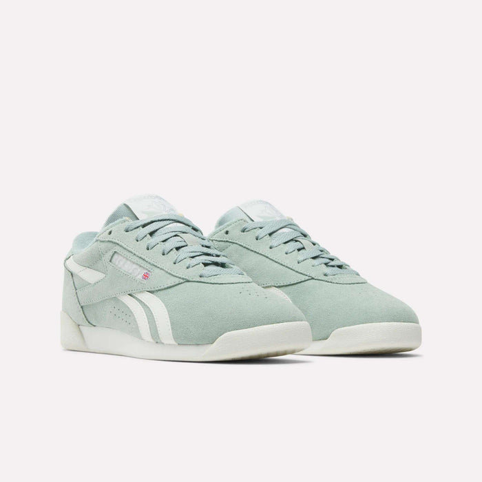 The Reebok Women's Freestyle Lo Shoes in light green suede feature white accents, soles, and side stripes, plus a subtle red logo tag near the laces. The retro sneakers are shown on a plain white background.
