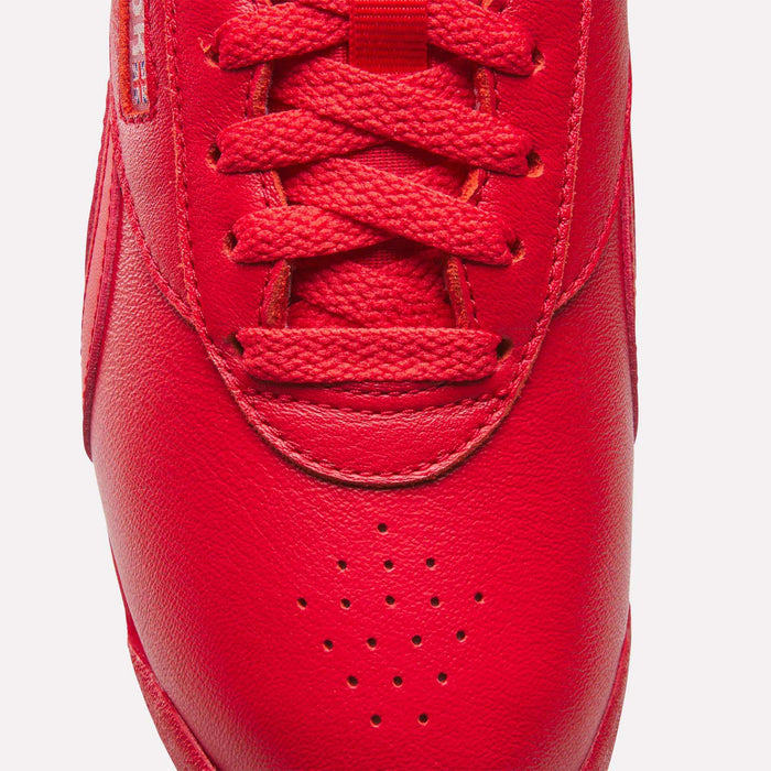 Close-up of the toe and laces of a red Reebok Women's Freestyle Lo Shoe, featuring a leather finish, red laces, toe perforations, and part of a fabric label on the side.