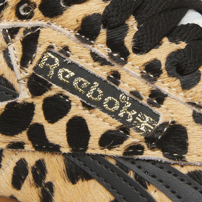 Close-up of Reebok Women's Freestyle Lo Shoes with a black and tan leopard print pony hair upper, stitched Reebok logo on a fabric tag, and black leather details inspired by classic '80s fitness trainer style.