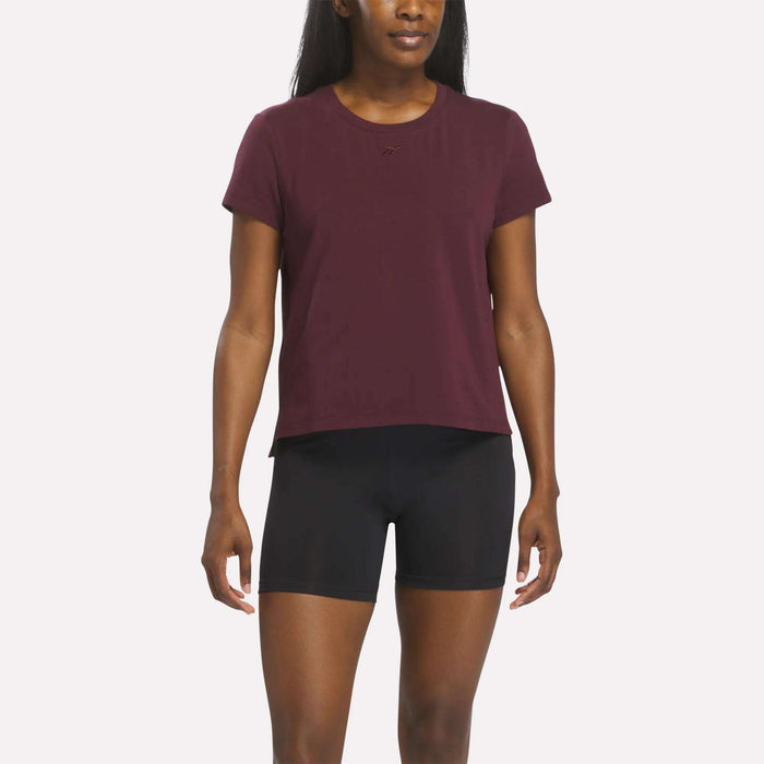 A woman wearing a Reebok Women's Graphic Glow Top in Wine Tasting and black fitted shorts stands against a plain white background.