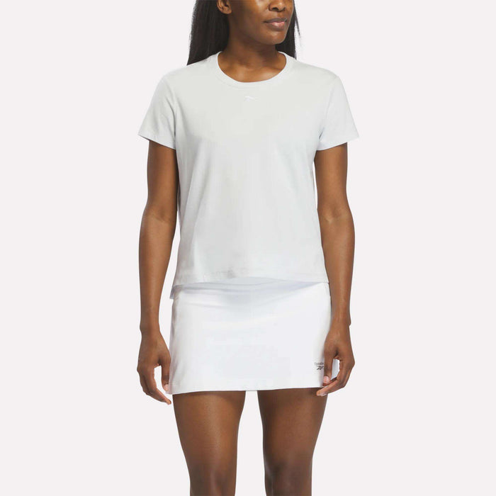 A woman stands facing forward with her arms relaxed at her sides, wearing a women's Reebok Graphic Glow Top in pearl grey and a white tennis skirt, set against a white background.