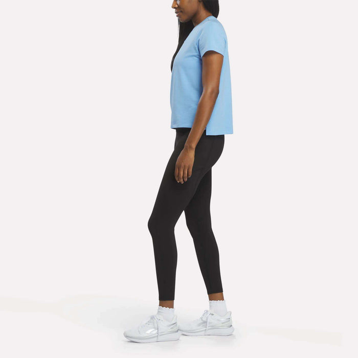 A woman in profile wears a light blue Women's Graphic Glow Top by Reebok, paired with black leggings and white sneakers, set against a plain white background.