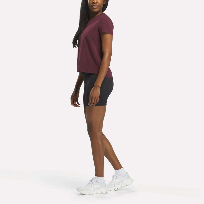 A woman stands sideways, wearing the Reebok Women's Graphic Glow Top in maroon, black fitted shorts, and white athletic shoes against a plain white background.