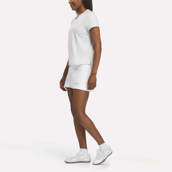 A woman faces left against a plain white background, wearing the Reebok Women's Graphic Glow Top with a matching skirt, paired with white athletic shoes and socks.