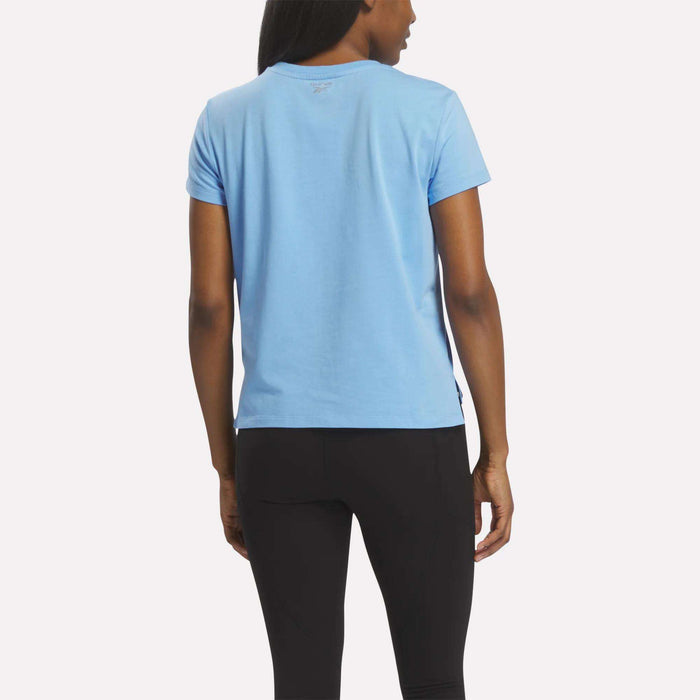 A woman stands facing away, wearing the Reebok Women's Graphic Glow Top in light blue with an embroidered logo and black pants, set against a plain light background.