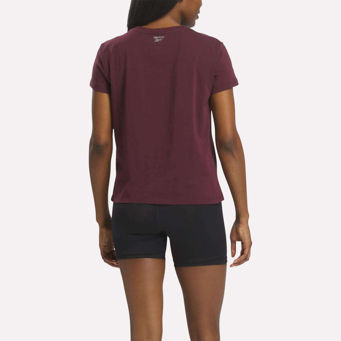 A person stands with their back to the camera, wearing the Reebok Women's Graphic Glow Top in maroon and black shorts with an embroidered logo, set against a plain white background.