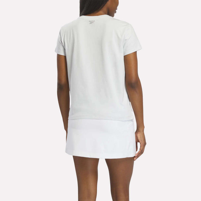 A woman faces away wearing the Reebok Women's Graphic Glow Top, a soft white short-sleeve tee, paired with a white skirt against a light background.