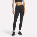 A person wears Reebok Women's Lux High-Rise Tights, a black sports bra, white socks, and black athletic shoes while standing against a plain light background.
