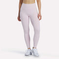 A woman wearing Reebok Women's High-Rise Leggings in Elemental Pink and white sneakers, standing against a plain white background. Her upper body and head are not visible.