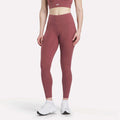 A woman models Reebok Women's High-Rise Tights in mauve with a matching sports bra and white sneakers, standing against a plain white background. Her head is cropped out of the image.