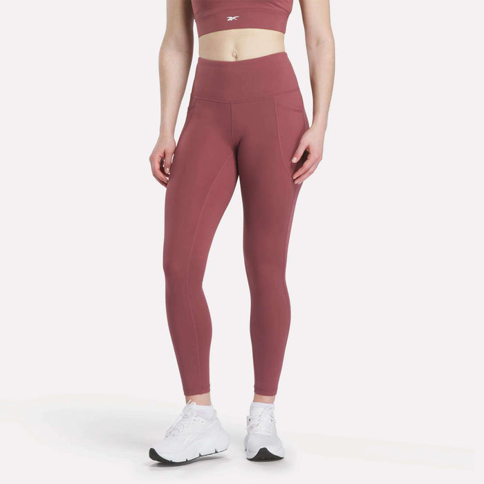 A woman models Reebok Women's High-Rise Tights in mauve with a matching sports bra and white sneakers, standing against a plain white background. Her head is cropped out of the image.