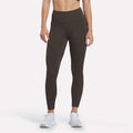 A person is shown from the neck down, standing against a plain white background and wearing Reebok Women’s High-Rise Leggings in Espresso along with white sneakers. The leggings are crafted with RBK-DRY performance fabric.