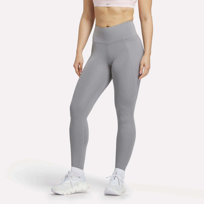 A woman models Reebok’s Lux-Lite High-Rise Tights with a light pink sports bra and white sneakers against a white background, highlighting an athletic, casual style.