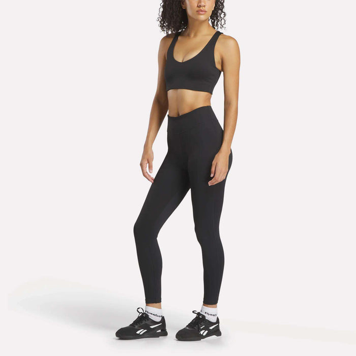 A woman faces forward in Reebok Women's Lux High-Rise Tights, a black sports bra, white socks, and black athletic shoes against a plain white background.
