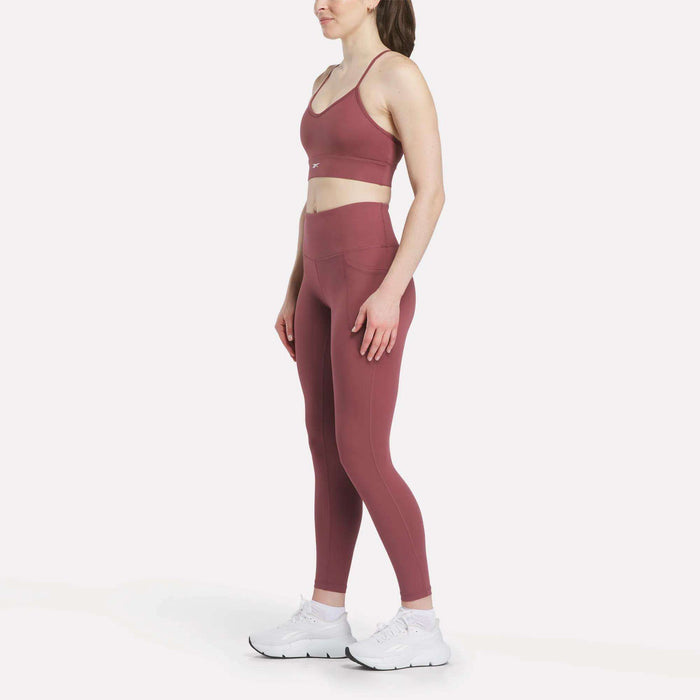 A woman in profile models Reebok Women's High-Rise Tights with a coordinating mauve sports bra and white athletic shoes, set against a plain white background.