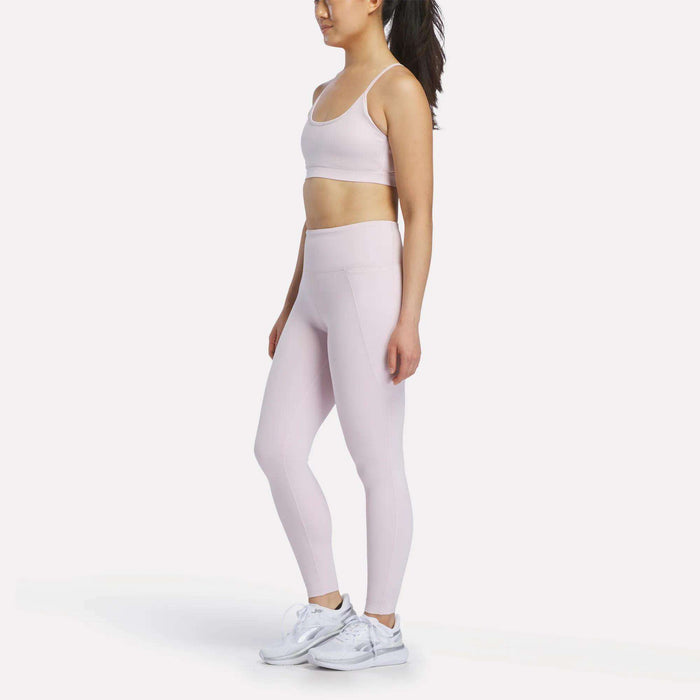 A woman stands sideways against a plain white background, wearing a light pink Reebok Women's High-Rise Leggings and matching sports bra set with white sneakers. Her hair is styled in a ponytail.