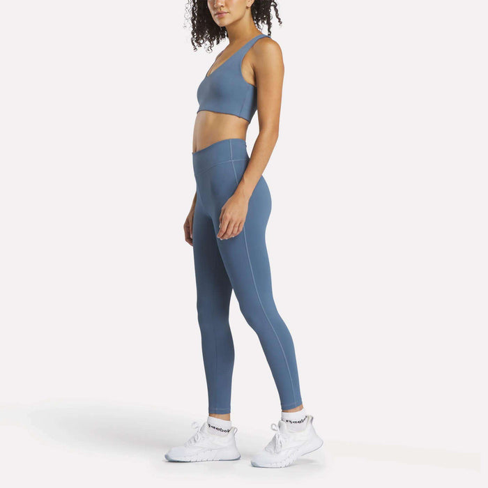 A woman stands in profile wearing Reebok's Women's Lux High-Rise Tights and a matching blue sports bra made from RBK-DRY fabric, paired with white athletic shoes, against a plain white background.