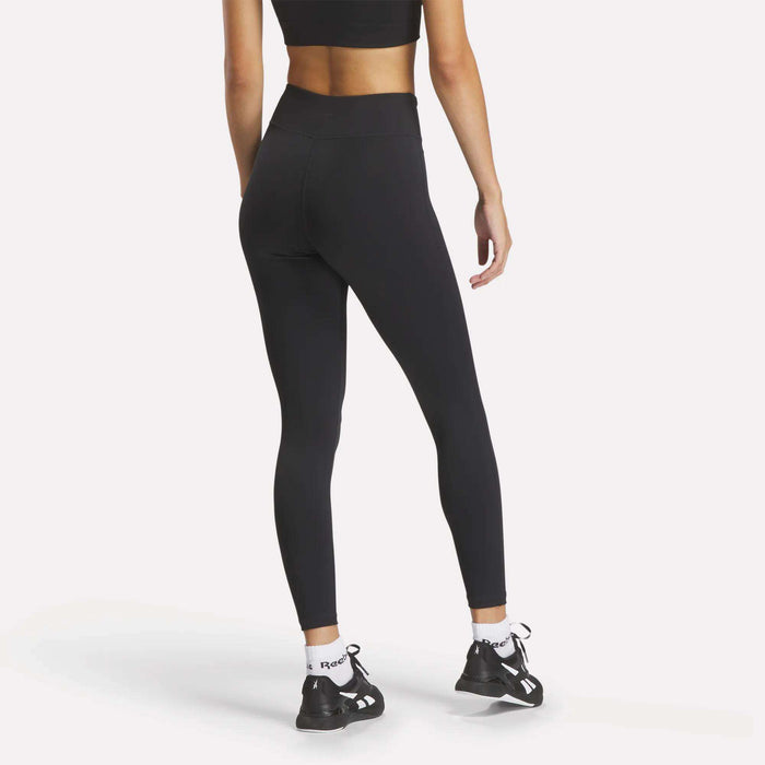 A woman, seen from behind, wears Reebok Women's Lux High-Rise Tights with a black sports bra, RBK-DRY socks, and black athletic shoes against a plain white background.