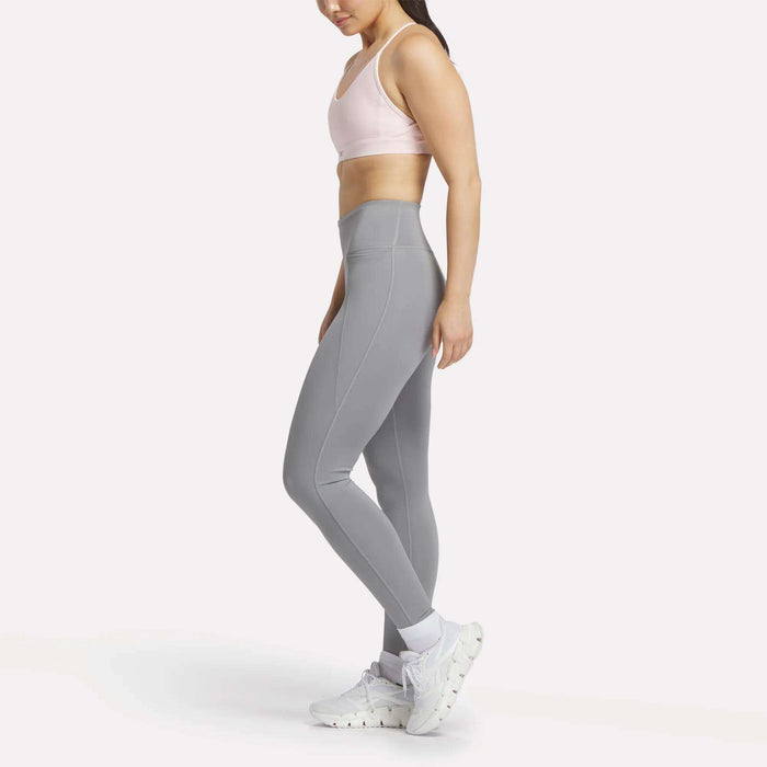 A woman stands sideways against a white background wearing Reebok Women's Lux-Lite High-Rise Tights, a light pink sports bra, white socks, and sneakers. The tights feature RBK-DRY performance fabric for comfort during training.