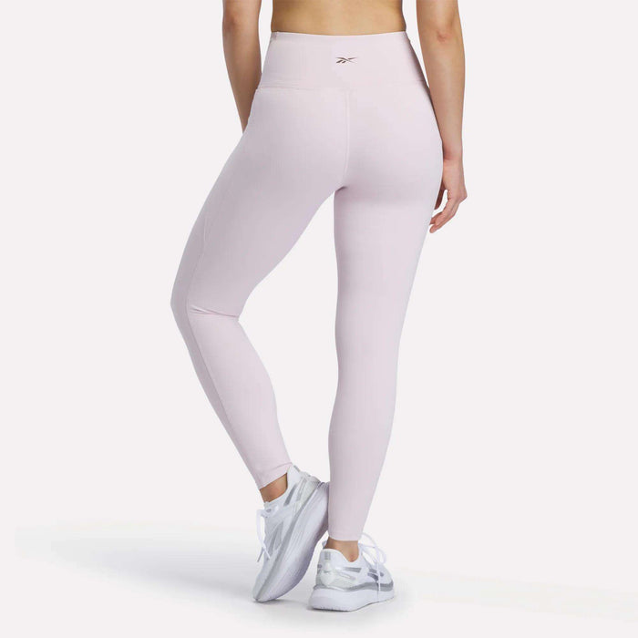 A person is wearing Reebok Women's High-Rise Leggings in light pink, which feature RBK-DRY technology, along with white athletic shoes, standing with their back to the camera against a plain white background.
