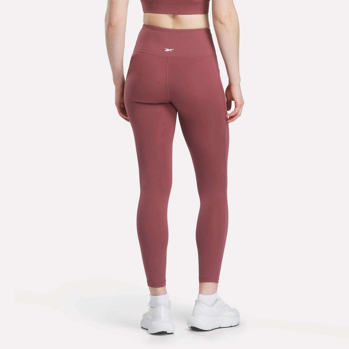 A person is seen from behind wearing Reebok Women's High-Rise Tights and a matching mauve sports bra with white athletic shoes, standing on a white surface.