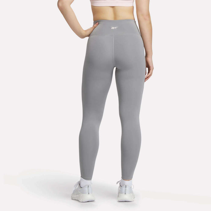 A woman stands facing away against a plain white background, wearing a light pink sports bra, white athletic shoes, and high-waisted gray Reebok Women's Lux-Lite High-Rise Tights.