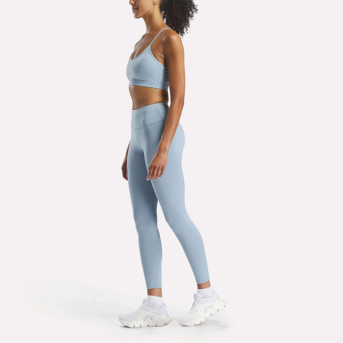 A woman in profile wears a light blue RBK-DRY sports bra, Reebok Women's Core High-Rise Leggings in matching blue, and white athletic shoes, standing against a plain white background.