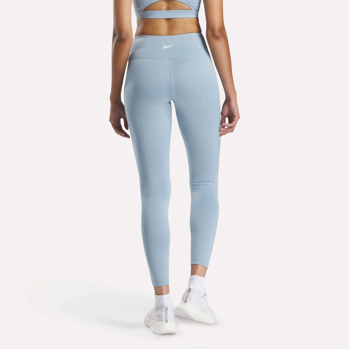 A woman wears Reebok Women's Core High-Rise Leggings and a matching light blue sports bra, both made with RBK-DRY fabric, standing against a white background with white sneakers, showing off the back of her outfit.