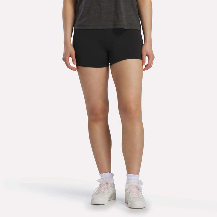A person is standing against a plain, light-colored background, wearing a dark t-shirt, white sneakers, and white ankle socks with ruffle trim. They are also wearing Reebok Women's High-Rise Mini Biker Shorts 3" in black. The person's face is not visible in the image.
