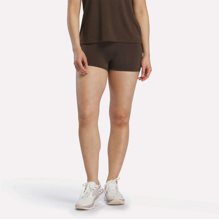 A person stands against a plain white background, wearing an espresso-colored Reebok Women's High-Rise Mini Biker Shorts 3" paired with a brown top and white sneakers. Their head is not visible in the image.
