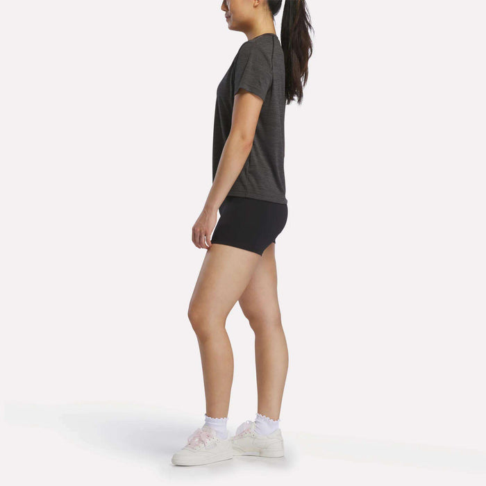 A woman stands in profile, facing left, wearing a dark t-shirt, Reebok Women's High-Rise Mini Biker Shorts 3", white frilled socks, and white sneakers. Her hair is tied back in a ponytail. The background is plain white.