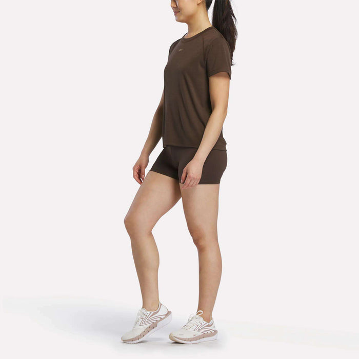 A woman with a ponytail stands sideways against a plain white background, wearing a brown t-shirt, white athletic shoes, and Reebok Women's High-Rise Mini Biker Shorts 3". The sweat-wicking training shorts from Reebok add comfort and style to her look.