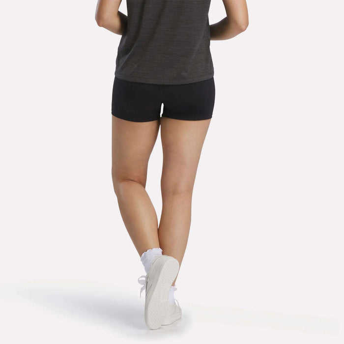 A person is shown from the back against a plain white background, standing with legs crossed and wearing a dark t-shirt, white socks, and white sneakers. They are also wearing black Reebok Women's High-Rise Mini Biker Shorts 3", which are designed with sweat-wicking fabric.