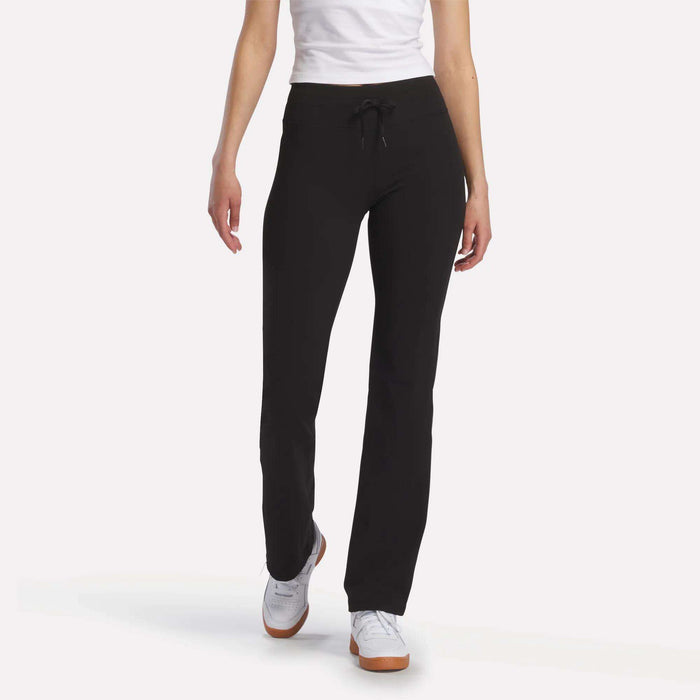A person stands against a white background, showcasing Reebok Women’s Highstreet High-Rise Pants in black, paired with a white shirt and white sneakers. The image is cropped at the shoulders to highlight the pants.