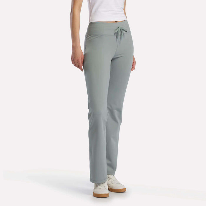 A person models Reebok Women's Highstreet High-Rise Pants in light gray, paired with a white fitted shirt and white sneakers, standing against a plain white background. The model's face is not shown.