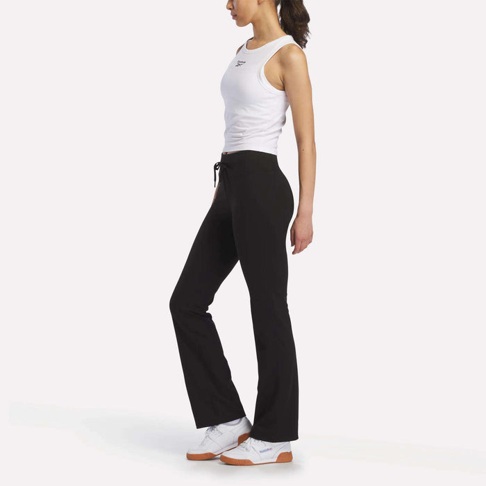 A person stands in side profile against a plain white background, wearing a white sleeveless top, Reebok Women's Highstreet High-Rise Pants in black, and white sneakers.