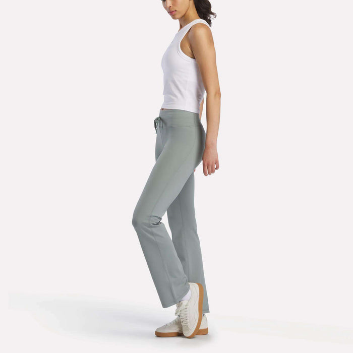 A woman stands sideways against a white background, wearing a white sleeveless top, Reebok Women's Highstreet High-Rise Pants in light gray-green with stretchy fabric, and white sneakers with beige soles.